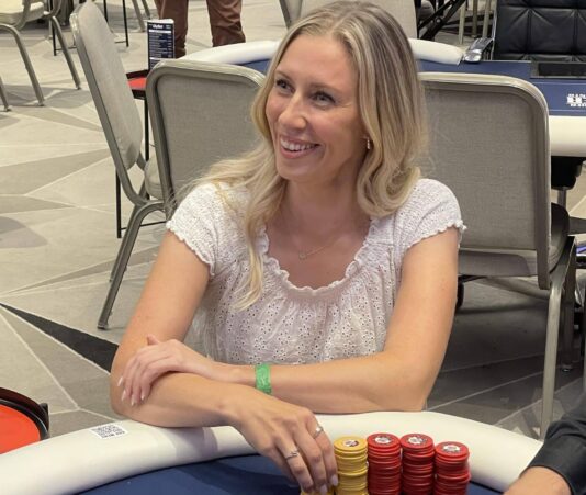 Linda Lahdenpää istuu pokeripöydän ääressä iloisesti hymyilleen valkoinen paita päällään ja kasa pokerimerkkejä edessään.