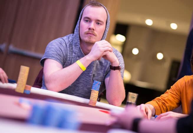 Mies istuu pokeripöydässä huppu päässä harmaassa hupparissa, etualalla on sinisiä pelimerkkejä ja oikealla oranssi hiha.