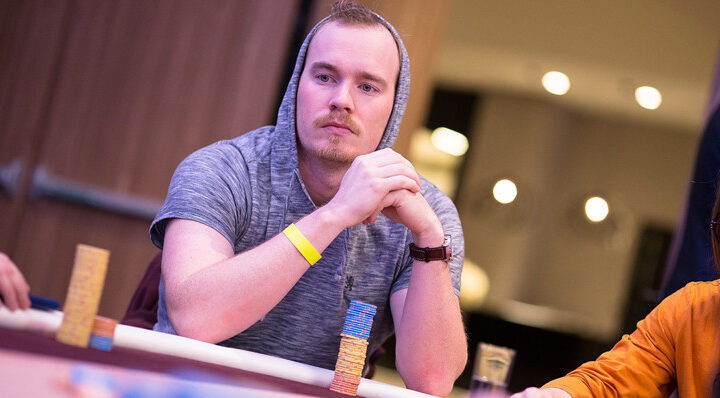 Mies istuu pokeripöydässä huppu päässä harmaassa hupparissa, etualalla on sinisiä pelimerkkejä ja oikealla oranssi hiha.