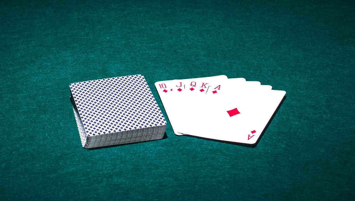 Vihreällä veralla oleva korttipakka ja vieressä viisi korttia käännettynä auki. Kortit ovat ruutua ja ne ovat arvoltaan kymppi, jätkä, rouva, kuningas ja ässä.