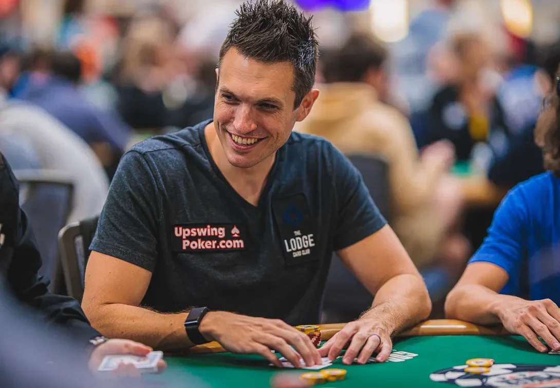 Doug Polk istuu vihreän pokeripöydän ääressä harmaassa T-paidassa ja pelimerkkejä ja pelikortteja edessään.