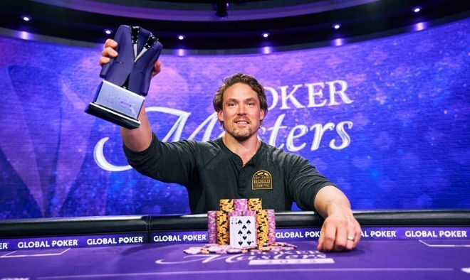 Mies istuu pokeripöydässä. Hänellä on kädessään voittopokaali, jota hän pitää ylhäällä. Edessään miehellä on pöydällä pelimerkkejä.