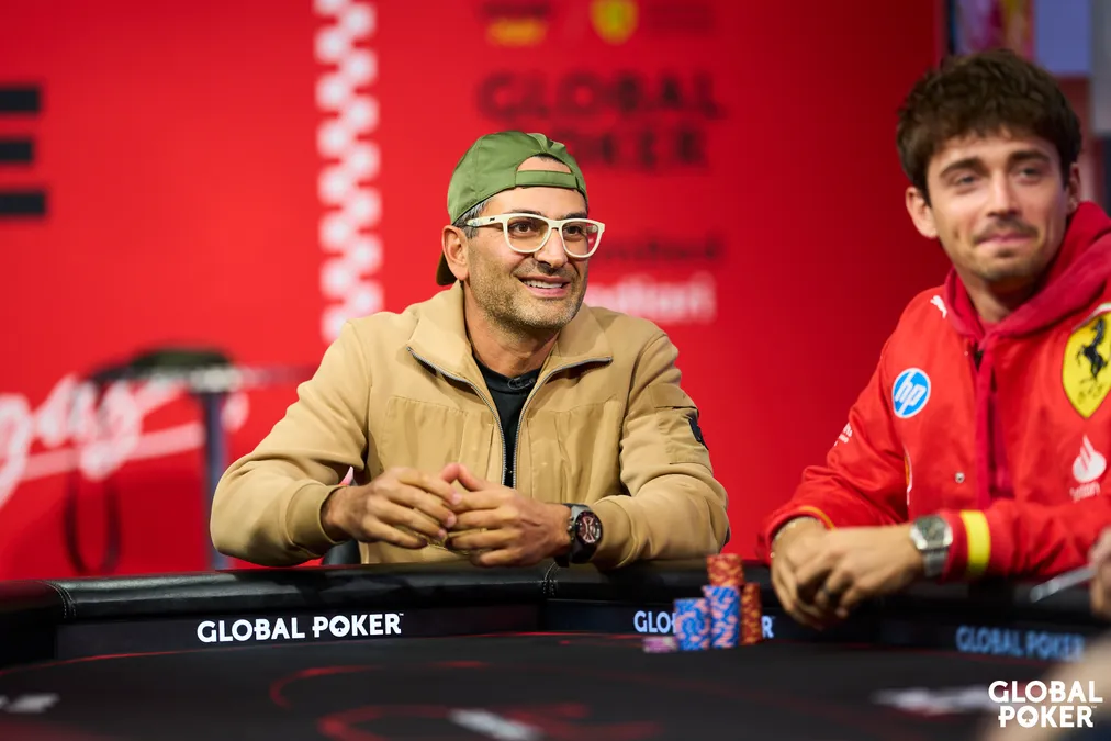 Kaksi miestä istuu pokeripöydän ääressä. Toisella on lippis väärin päin päässä ja beige takki. Toisella on mustat hiukset ja ferrarin punaiset vaatteet.
