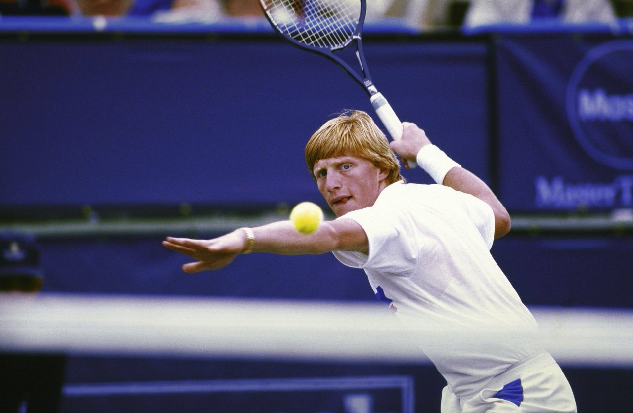 Mies pelaamassa tennistä. Miehellä on oranssi tukka ja valkea paita. Hän katsoo keskittyneenä palloon.