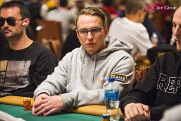 Benjamin Rolle harmaassa hupparissa istuu pokeripöydän ääressä. Pöydällä on vesipullo. Taustalla näkyy muita ihmisiä. Miehellä on silmälasit