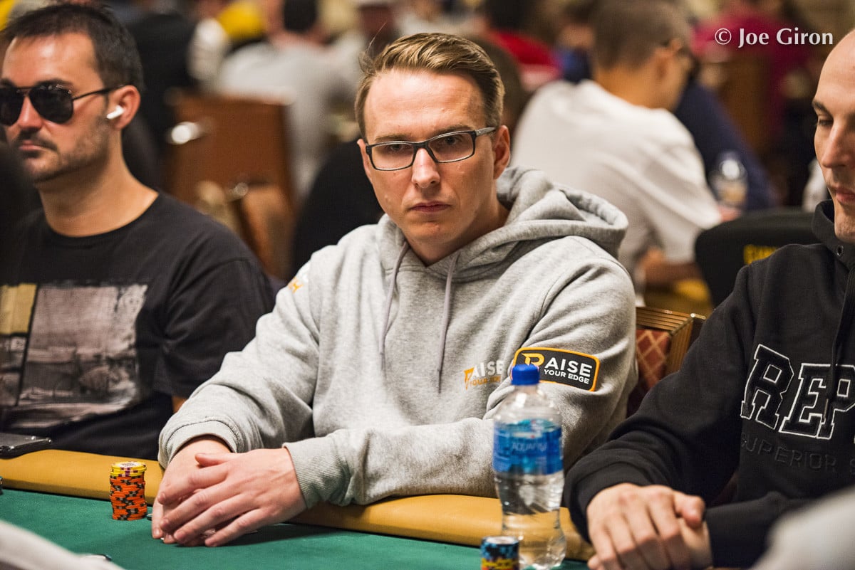 Mies harmaassa hupparissa istuu pokeripöydän ääressä. Pöydällä on vesipullo. Taustalla näkyy muita ihmisiä. Miehellä on silmälasit