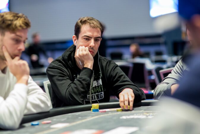 Mietteliään näköinen mies istuu pokeripöydän ääressä ja nojaa käteensä. Miehellä on musta huppari ja nappikuulokkeet korvissa.