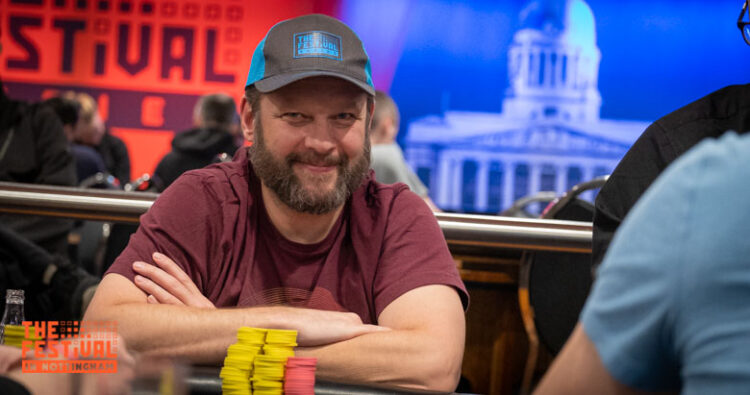 Mies istuu pokeripöydän ääressä. Pöydällä edessä on pelimerkkejä. Miehellä on parta ja päässään lippahattu. Miehen paita on tumman punainen.