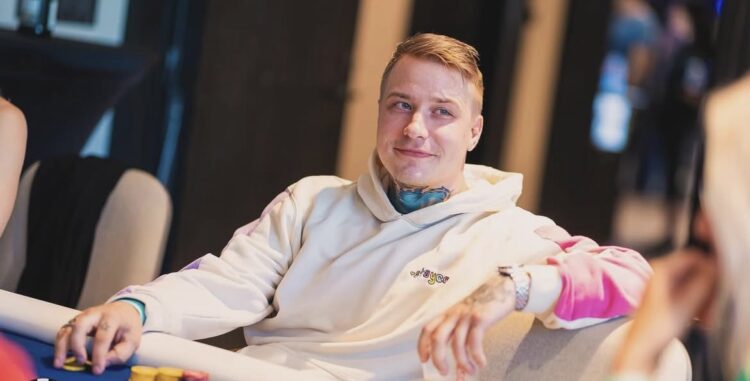 Mies istuu pokeripöydässä päällään vaalea huppari.Toisella kädellä mies nojaa tuoliin ja toisessa kädessä hänellä on pelimerkkejä.