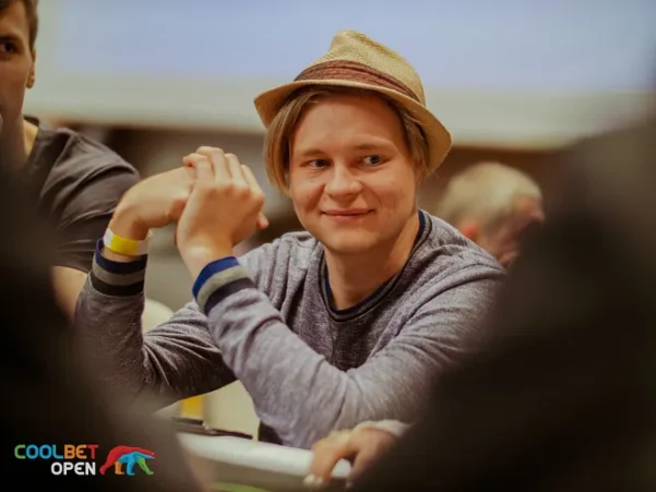 Mies istuu pokeripöydässä ja hymyilee. Hänelle on päässään vaaleanruskea lierihattu ja päällään vaaleansininen paita.