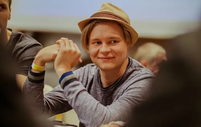 Mies istuu pokeripöydässä ja hymyilee. Hänelle on päässään vaaleanruskea lierihattu ja päällään vaaleansininen paita.