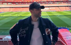 Mies seisoo katsomossa. Takana näkyy nurmikenttä sekä vastapäinen katsomo. Miehellä on päällään musta takki ja musta lippis.