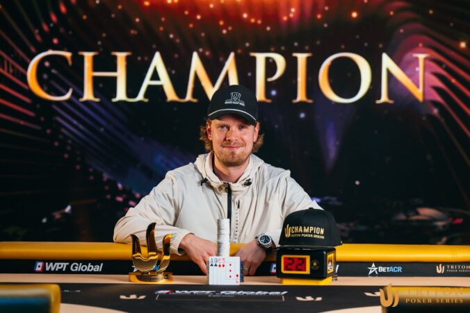 Mies poseeraa pokeripöydän takana. Pöydällä on pelimerkkejä, kortteja. lippis ja pokaali. Miehellä on päässään musta lippahattu.