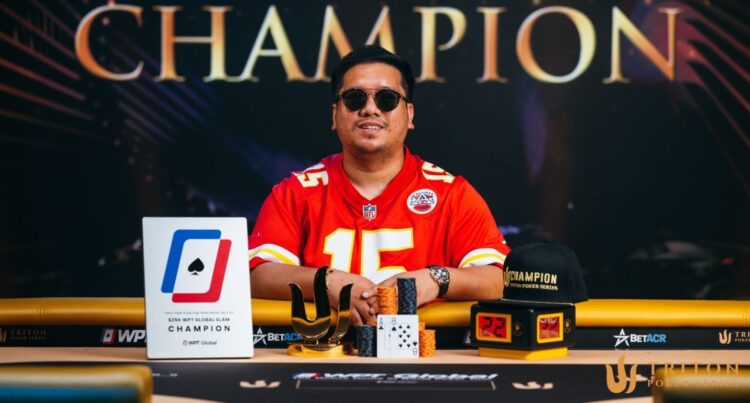 Mies punaisessa paidassa poseeraa pokeripöydän edessä. Pöydällä on pelikortteja ja pelimerkkejä. Taustalla on kultaista tekstiä. Miehellä on aurinkolasit.