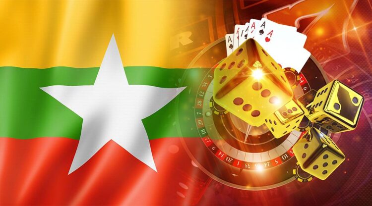 Digitaalinen kuvakollaasi, joka yhdistää Myanmarin lipun ja kasinopelaamisen symboliikkaa. Kuvan taustaa ja vasenta laitaa hallitsee suuri, silkkisen näköisenä aaltoileva Myanmarin lippu, jossa on kelta-viher-punaiset vaakaraidat ja suuri valkoinen tähti keskellä. Lipun päälle on sommiteltu kokoelma rahapelaamiseen liittyviä elementtejä. Näihin kuuluvat muun muassa rulettipyörä, useita suuria, kultaisia ja läpikuultavia noppia, neljän ässän korttikäsi sekä pelimerkkejä. Kuva on valaistu kirkkailla, lämpimillä valoilla ja heijastuksilla, mikä luo energisen ja viihdekeskeisen tunnelman