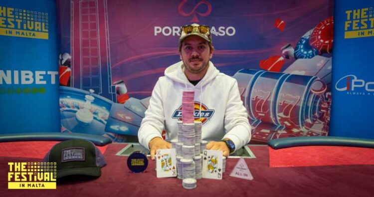 Mies valkeassa hupparissa istuu pokeripöydän takana. Pöydällä on pelimerkeistä kasattu torni. Pöytä on punainen ja tausta värikäs.