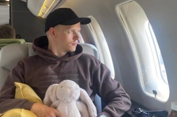 Mies istuu lentokoneessa. Hänellä on sylissään pehmolelu, joka esittää pupua.Miehellä on päällää harmaa huppari ja tumma lippis.