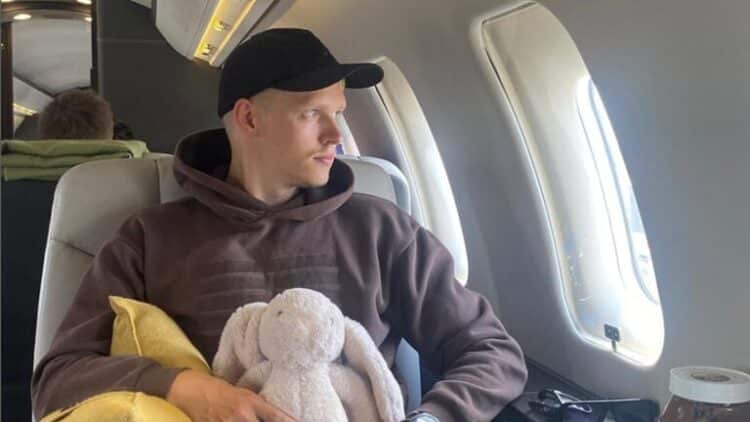 Mies istuu lentokoneessa. Hänellä on sylissään pehmolelu, joka esittää pupua.Miehellä on päällää harmaa huppari ja tumma lippis.