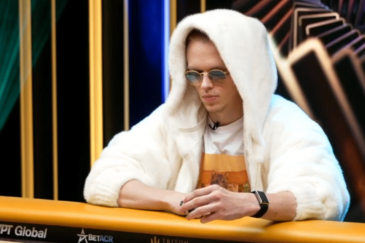 Mies istuu pokeripöydän äärellä valkoisessa hupparissa. Miehellä on päässään pyöreät auronkolasit ja edessään pelimerkkejä.