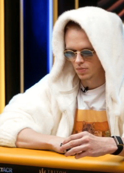 Mies istuu pokeripöydän äärellä valkoisessa hupparissa. Miehellä on päässään pyöreät auronkolasit ja edessään pelimerkkejä.