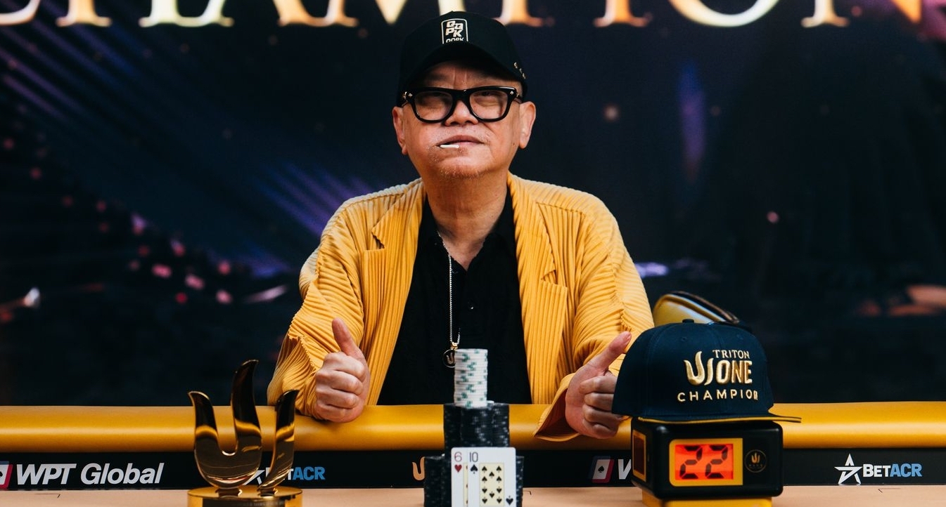 Mies istuu pokeripöytän ääressä. Miehellä on musta lippis ja keltainen takki. Mies näyttää molempia peukaloitaan ylöspäin.