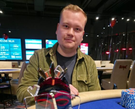 Mies poseeraa palkintopokaalin kanssa pokeripöydän ääressä. Sinisellä pöydällä on myös pelimerkkejä.