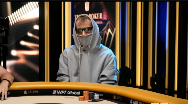Mies verhoutuneena huppariin istuu pokeripöydässä. Huppari on vaalean harmaa ja miehellä on aurinkolasit.