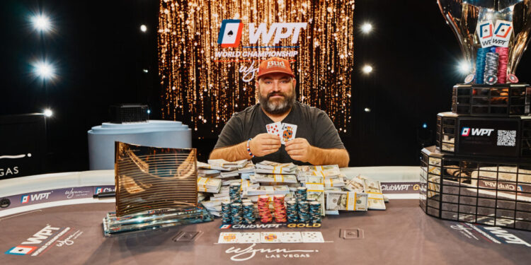 Mies istuu pokeripöydässä. Hän pitelee kädessään pelikortteja. Miehen edessä pöydällä on iso kasa rahaa ja pelimerkkejä.