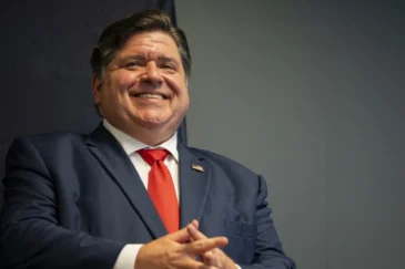 Illinoisin kuvernööri Pritzker sinisessä puvussa, punaisessa solmiossa, kädet ristittynä