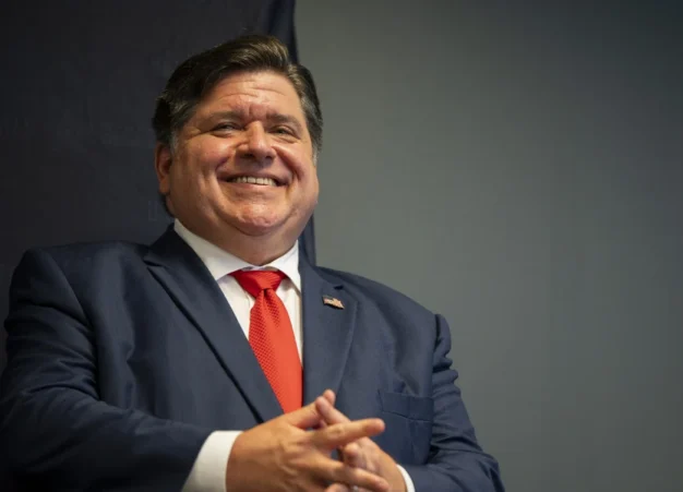 Illinoisin kuvernööri Pritzker sinisessä puvussa, punaisessa solmiossa, kädet ristittynä