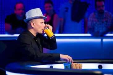 Ilaei Sahamies istuu sinisävyisessä pokeripöydässä hattu päässään, juoden lasista appelsiinimehua