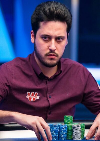 Adrian Mateos istuu pokeripöydässä. Hänellä on päällään punainen kauluspaita. Hänen kädet ovat pöydällä, jossa on myös pelimerkkejä.