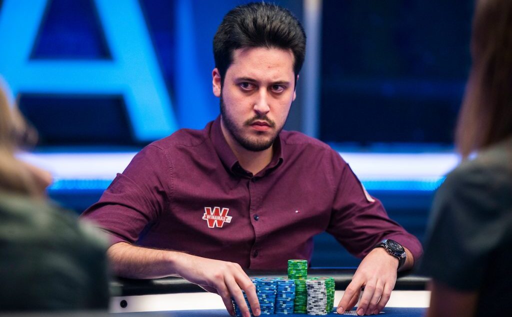 Adrian Mateos istuu pokeripöydässä. Hänellä on päällään punainen kauluspaita. Hänen kädet ovat pöydällä, jossa on myös pelimerkkejä.