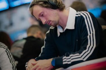 Mies istuu pokeripöydässä. Hänellä päällään pitkähihainen kauluspaita, jonka hihoissa on raitoja. Päässä miehellä on kuulokkeet.