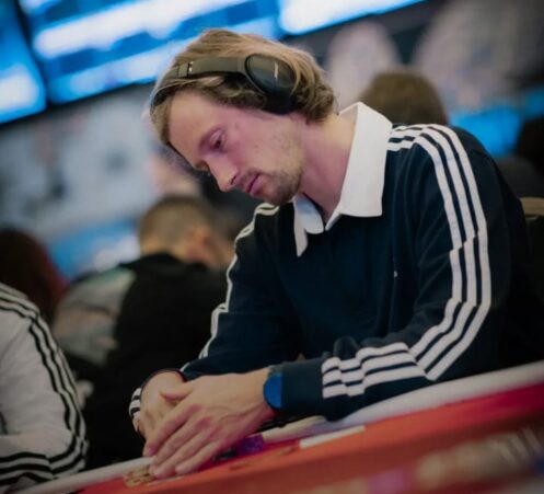 Mies istuu pokeripöydässä. Hänellä päällään pitkähihainen kauluspaita, jonka hihoissa on raitoja. Päässä miehellä on kuulokkeet.