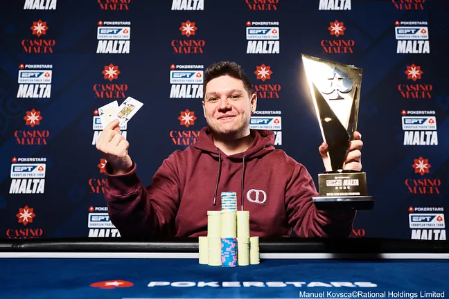 Kuvassa on tyytyväisen näköinen mies, joka juhlii voittoaan PokerStarsin European Poker Tour (EPT) -turnauksessa Maltalla. Hän istuu sinisen pelipöydän ääressä ja hymyilee leveästi. Voittaja pitelee toisessa kädessään suurta kultaista pokaalia ja esittelee toisessa kädessään ylpeänä voittokättään: kahta ässää. Hänen edessään on korkea pino turnausmerkkejä. Taustalla oleva sininen mainosseinä vahvistaa tapahtuman arvokkuuden. Kuva on klassinen voittajan muotokuva, joka on täynnä iloa ja ylpeyttä merkittävän saavutuksen jälkeen. Valokuvan on ottanut Manuel Kovsca.