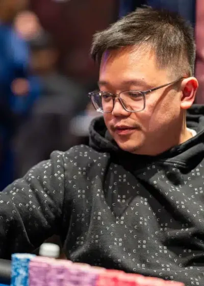 Kuvassa on aasialainen mies, joka on syvästi keskittynyt pokeriturnaukseen. Hän istuu kirkkaansinisen pelipöydän ääressä pukeutuneena mustaan kuviolliseen huppariin ja silmälaseihin. Hänen edessään pöydällä on erittäin suuri ja vaikuttava pino pelimerkkejä, jotka on järjestetty siisteihin, pitkiin riveihin pääasiassa purppuran- ja punaisen värisinä. Miehen oikea käsi on juuri kurkottamassa kohti merkkejä, valmiina tekemään panostuksen. Hänen ilmeensä on analyyttinen ja rauhallinen, ja hänen katseensa on suunnattu tiukasti peliin. Tausta on sumea, korostaen pelaajan intensiivistä keskittymistä hetkeen.