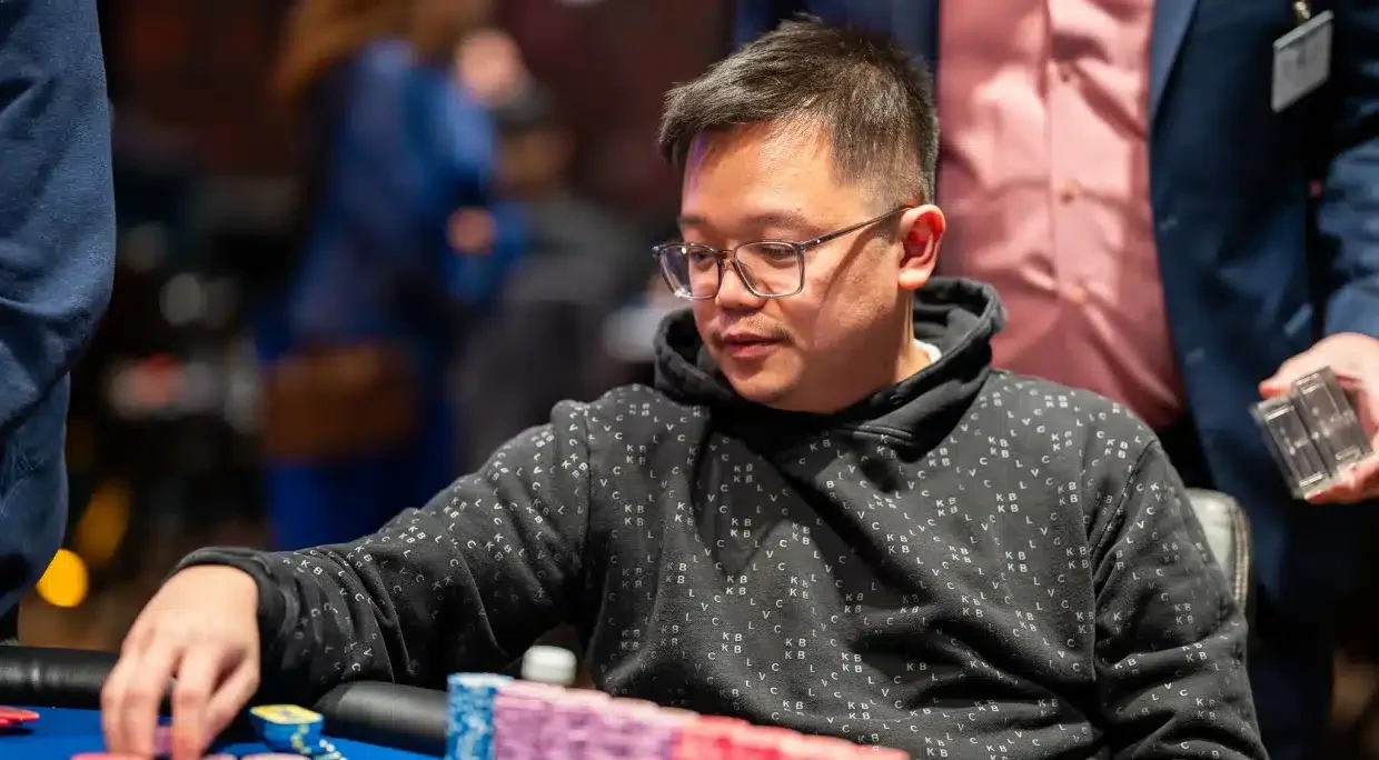 Kuvassa on aasialainen mies, joka on syvästi keskittynyt pokeriturnaukseen. Hän istuu kirkkaansinisen pelipöydän ääressä pukeutuneena mustaan kuviolliseen huppariin ja silmälaseihin. Hänen edessään pöydällä on erittäin suuri ja vaikuttava pino pelimerkkejä, jotka on järjestetty siisteihin, pitkiin riveihin pääasiassa purppuran- ja punaisen värisinä. Miehen oikea käsi on juuri kurkottamassa kohti merkkejä, valmiina tekemään panostuksen. Hänen ilmeensä on analyyttinen ja rauhallinen, ja hänen katseensa on suunnattu tiukasti peliin. Tausta on sumea, korostaen pelaajan intensiivistä keskittymistä hetkeen.