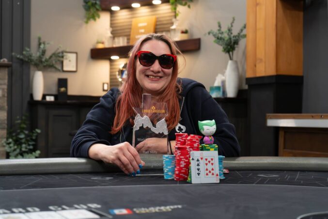 Kuvassa on onnellinen nainen, joka juhlii voittoaan pokeriturnauksessa. Hän hymyilee leveästi kameralle punaiseksi värjätyissä hiuksissaan ja suurissa aurinkolaseissaan. Voittaja istuu pelipöydän takana ja pitelee kädessään läpinäkyvää pokaalia, jossa lukee "Lodge Card Club Champion". Hänen edessään on suuret pinot pelimerkkejä, voittokäsi (ässä ja kymppi) sekä pieni henkilökohtainen maskottihahmo. Tausta on tyylikäs ja rauhallinen, mikä viittaa laadukkaaseen pokeriklubiin. Koko kuvasta välittyy aito voitonriemu ja onnistumisen tunne.