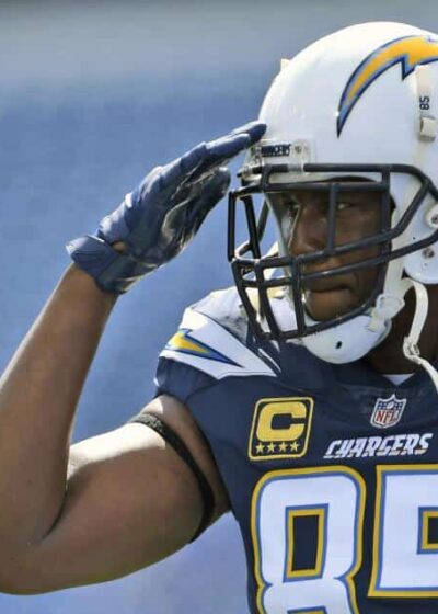 Kuvassa on NFL-legenda Antonio Gates, yksi kaikkien aikojen parhaista tight endeistä. Hän seisoo amerikkalaisen jalkapallon kentällä pukeutuneena San Diego Chargersin laivastonsiniseen pelipaitaan, jossa on numero 85 ja kapteenin "C"-merkki. Gates on kohottanut oikean kätensä kypäränsä reunalle kunnioittavaan sotilastervehdykseen. Hänen kypärässään on Chargersin ikoninen salama-logo. Gatesin katse on vakava ja keskittynyt, ja koko ele huokuu kunnioitusta. Tausta on sumennettu, mikä kohdistaa kaiken huomion tähän ikoniseen pelaajaan ja hänen voimakkaaseen eleeseensä.