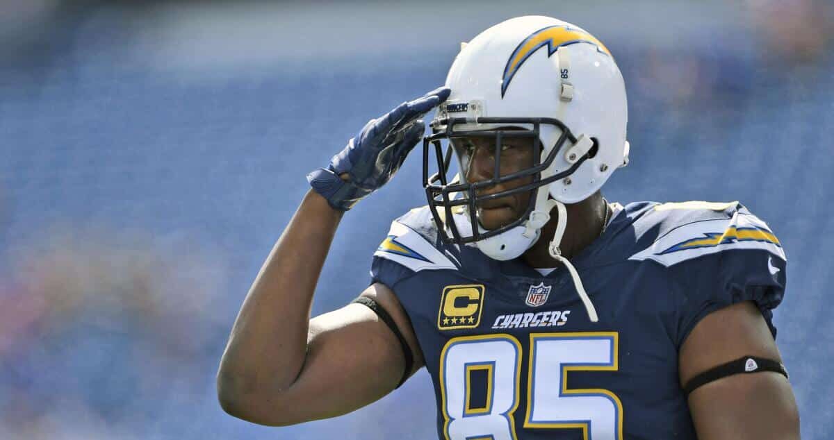 Kuvassa on NFL-legenda Antonio Gates, yksi kaikkien aikojen parhaista tight endeistä. Hän seisoo amerikkalaisen jalkapallon kentällä pukeutuneena San Diego Chargersin laivastonsiniseen pelipaitaan, jossa on numero 85 ja kapteenin "C"-merkki. Gates on kohottanut oikean kätensä kypäränsä reunalle kunnioittavaan sotilastervehdykseen. Hänen kypärässään on Chargersin ikoninen salama-logo. Gatesin katse on vakava ja keskittynyt, ja koko ele huokuu kunnioitusta. Tausta on sumennettu, mikä kohdistaa kaiken huomion tähän ikoniseen pelaajaan ja hänen voimakkaaseen eleeseensä.