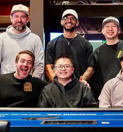 Kuvassa on kuuden miehen iloinen ryhmä poseeraamassa yhdessä kirkkaansinisen pokeripöydän ääressä. Kolme miehistä istuu etualalla ja kolme seisoo heidän takanaan. Kaikki hymyilevät leveästi kameralle, mutta erityisesti eturivin vasemmassa reunassa oleva mies tuulettaa erittäin innostuneesti kädet levällään ja suu auki. Ryhmällä on yllään rentoja vaatteita, kuten huppareita ja lippalakkeja, ja he vaikuttavat olevan ystäviä tai pelikumppaneita. Tausta on moderni pokerihuone. Kuvan tunnelma on toverillinen, positiivinen ja täynnä hyvää energiaa.