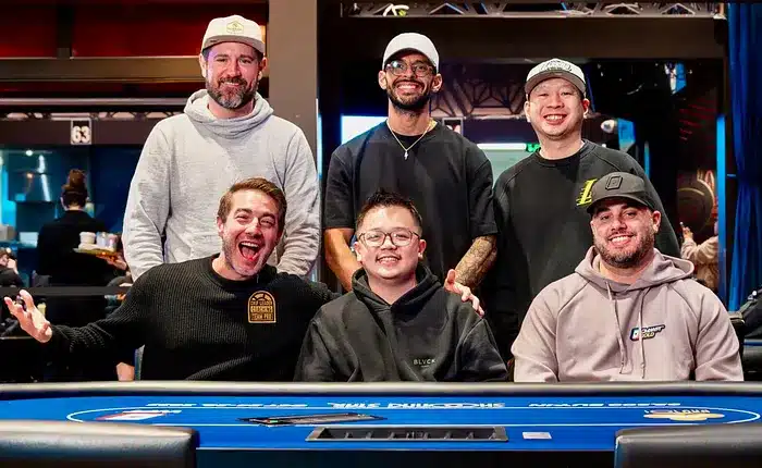 Kuvassa on kuuden miehen iloinen ryhmä poseeraamassa yhdessä kirkkaansinisen pokeripöydän ääressä. Kolme miehistä istuu etualalla ja kolme seisoo heidän takanaan. Kaikki hymyilevät leveästi kameralle, mutta erityisesti eturivin vasemmassa reunassa oleva mies tuulettaa erittäin innostuneesti kädet levällään ja suu auki. Ryhmällä on yllään rentoja vaatteita, kuten huppareita ja lippalakkeja, ja he vaikuttavat olevan ystäviä tai pelikumppaneita. Tausta on moderni pokerihuone. Kuvan tunnelma on toverillinen, positiivinen ja täynnä hyvää energiaa.