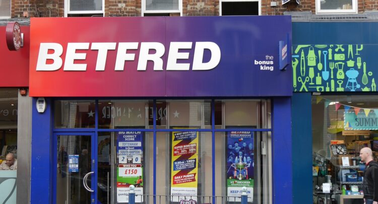 Kuvassa on brittiläisen Betfred-vedonlyöntiliikkeen katutason myymälän julkisivu, joka on otettu suoraan kadulta. Liikkeen suuri kyltti on huomiota herättävä, ja sen väri liukuu punaisesta tummansiniseen. Kyltissä lukee valkoisilla suuraakkosilla "BETFRED" sekä slogan "the bonus king". Koko julkisivu on kehystetty kirkkaansinisellä. Suurissa ikkunoissa on useita värikkäitä mainosjulisteita, jotka mainostavat jalkapallovetoja ja muita bonuksia. Liike sijaitsee vanhemman tiilirakennuksen pohjakerroksessa. Oikealla puolella on toinen liike, jonka ohi kävelee kaljupäinen mies kantaen valkoista muovikassia.