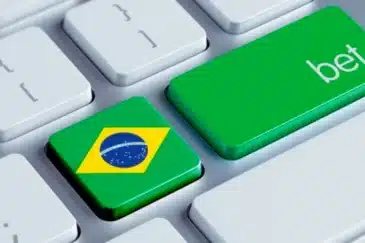 Kuvassa on tyylitelty lähikuva modernista tietokoneen näppäimistöstä. Kuva on sommitelma, joka kuvaa online-vedonlyöntiä Brasiliassa. Kaksi näppäintä on nostettu esiin: Enter-näppäin on korvattu suurella vihreällä näppäimellä, jossa lukee sana "bet" (lyö vetoa). Heti sen vieressä on näppäin, johon on kuvattu Brasilian lippu. Yksinkertainen mutta tehokas visuaalinen sommittelu viestii selkeästi brasilialaisille suunnatuista tai Brasiliassa tapahtuvasta nettipelaamisesta ja -vedonlyönnistä. Se on tyypillinen kuvituskuva, jota käytetään aiheeseen liittyvissä uutisissa ja artikkeleissa.