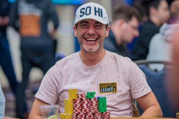 Kuvassa on yhdysvaltalainen pokeriammattilainen Chance Kornuth, joka on vangittu iloiseen ja rentoon hetkeen kesken turnauksen. Hän nauraa leveästi ja vapautuneesti kameralle. Kornuthilla on yllään vaalea t-paita ja valkoinen lippalakki, jossa lukee "SO HI". Hänen edessään vihreällä pelipöydällä on valtava pino erivärisiä pelimerkkejä, ja hänen paitaansa on kiinnitetty keltainen "CHIP LEADER" -merkki, mikä osoittaa hänen olevan turnauksen johdossa. Kuva on täynnä positiivista energiaa ja itseluottamusta, ja sen on ottanut valokuvaaja Hayley Hochstetler.