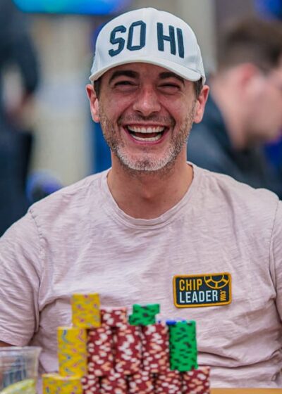 Kuvassa on yhdysvaltalainen pokeriammattilainen Chance Kornuth, joka on vangittu iloiseen ja rentoon hetkeen kesken turnauksen. Hän nauraa leveästi ja vapautuneesti kameralle. Kornuthilla on yllään vaalea t-paita ja valkoinen lippalakki, jossa lukee "SO HI". Hänen edessään vihreällä pelipöydällä on valtava pino erivärisiä pelimerkkejä, ja hänen paitaansa on kiinnitetty keltainen "CHIP LEADER" -merkki, mikä osoittaa hänen olevan turnauksen johdossa. Kuva on täynnä positiivista energiaa ja itseluottamusta, ja sen on ottanut valokuvaaja Hayley Hochstetler.