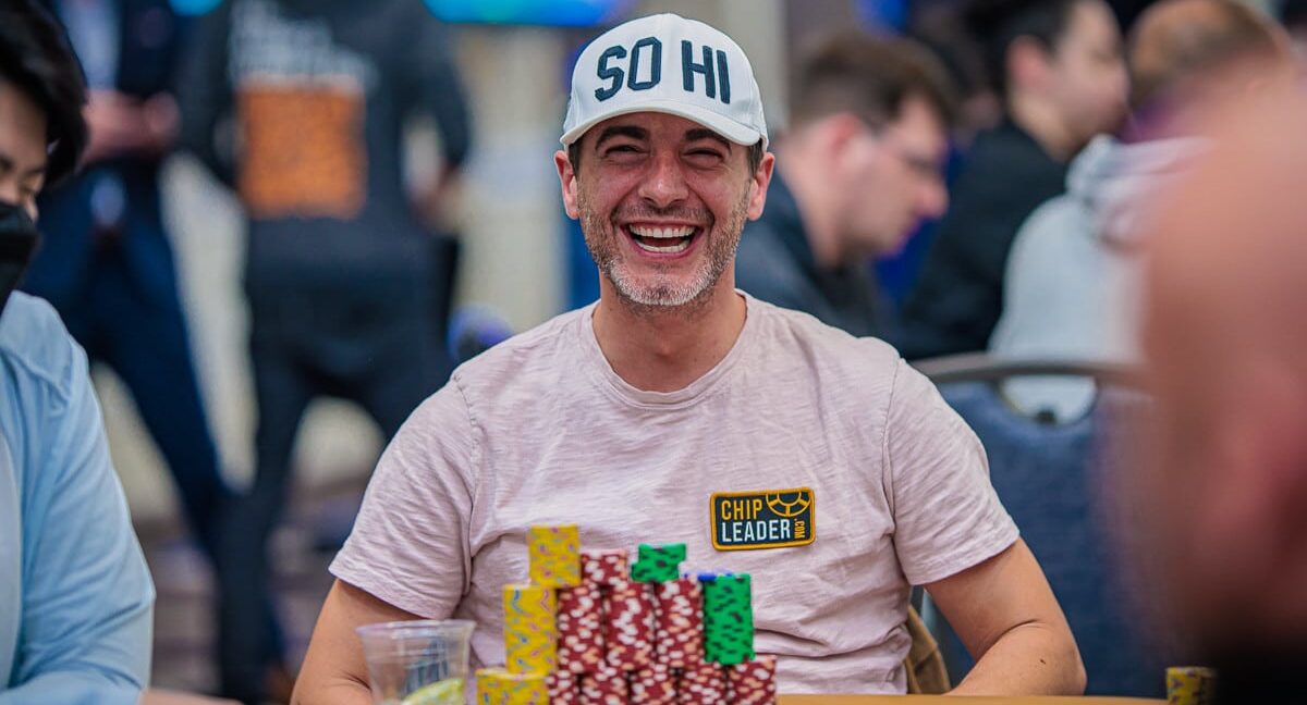 Kuvassa on yhdysvaltalainen pokeriammattilainen Chance Kornuth, joka on vangittu iloiseen ja rentoon hetkeen kesken turnauksen. Hän nauraa leveästi ja vapautuneesti kameralle. Kornuthilla on yllään vaalea t-paita ja valkoinen lippalakki, jossa lukee "SO HI". Hänen edessään vihreällä pelipöydällä on valtava pino erivärisiä pelimerkkejä, ja hänen paitaansa on kiinnitetty keltainen "CHIP LEADER" -merkki, mikä osoittaa hänen olevan turnauksen johdossa. Kuva on täynnä positiivista energiaa ja itseluottamusta, ja sen on ottanut valokuvaaja Hayley Hochstetler.
