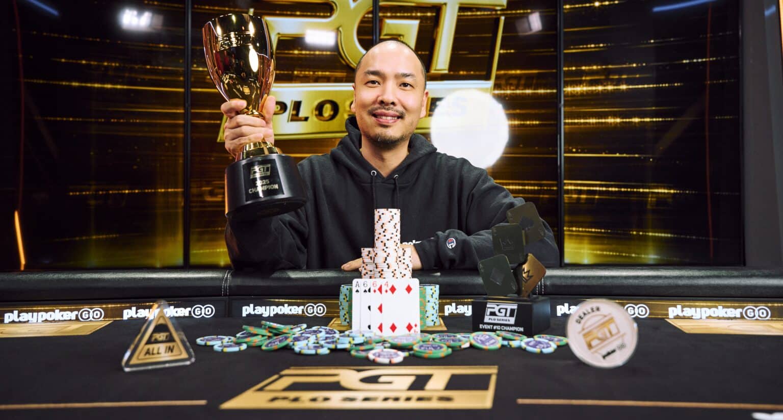 Kuvassa on iloisesti hymyilevä, kaljuuntuva mies, joka juhlii voittoaan PokerGO Tour (PGT) -turnauksessa. Hänellä on yllään musta huppari, ja hän pitelee ylpeänä toisessa kädessään suurta kultaista voittopokaalia. Hänen edessään mustalla pelipöydällä on korkea pino pelimerkkejä, joiden eteen on aseteltu voittokäsi: neljä ässää. Pöydällä on myös useita muita pienempiä PGT-palkintoja, kuten "Event #8 Champion" -laatta. Taustalla loistaa suuri, kulta-musta digitaalinen näyttö, jossa näkyy PGT-logo ja teksti "PLO SERIES". Kuva on täynnä tyytyväisyyttä ja menestyksen tuntua.