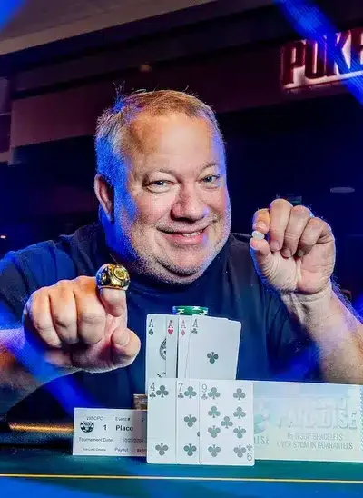 Kuvassa on riemukkaasti hymyilevä mies, joka poseeraa pokeriturnauksen voittajana. Hän istuu kirkkaansinisen pelipöydän ääressä ja ojentaa toista kättään ylpeänä eteenpäin, esitellen suurta kultaista mestaruussormusta sormessaan. Hänen edessään pöydällä on voittokäsi – neljä ässää – sekä ensimmäisen sijan palkintolaatta ja suuri "Ticket to Paradise" -voittokuponki. Taustalla näkyy "POKER ROOM" -kyltti, ja kuvaan luovat dramaattisen efektin kirkkaat siniset valot, jotka muodostavat ristikkäisen kuvion. Miehen ilme on täynnä aitoa iloa ja tyytyväisyyttä.